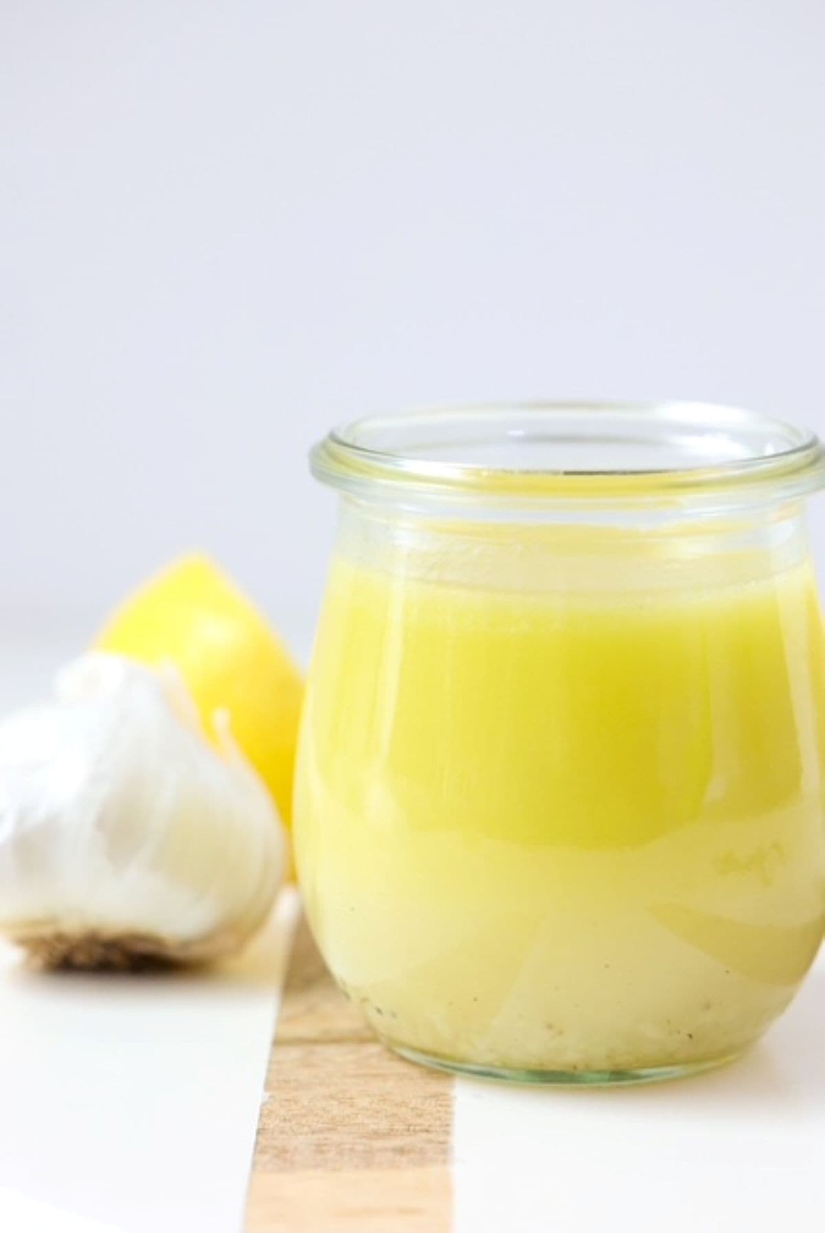 Lemon Garlic Salad Dressing on a white board with a wooden stripe. One garlic bulb and lemon sit to the back right.