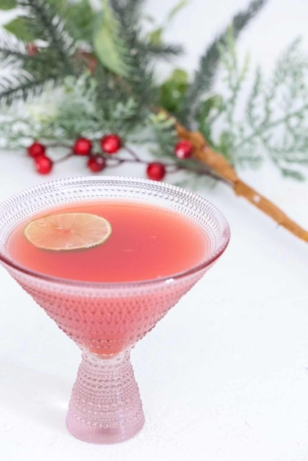 One decorative martini style glass with Christmas punch topped with a slice of lime. A Christmas tree branch is in the background.