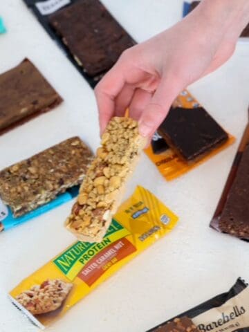 A child's hand picking up a Nature Valley Protein Bar with other protein bars in the background.