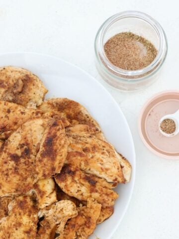 Seasoned chicken on a white plate with chicken seasoning in a n open jar to the side.