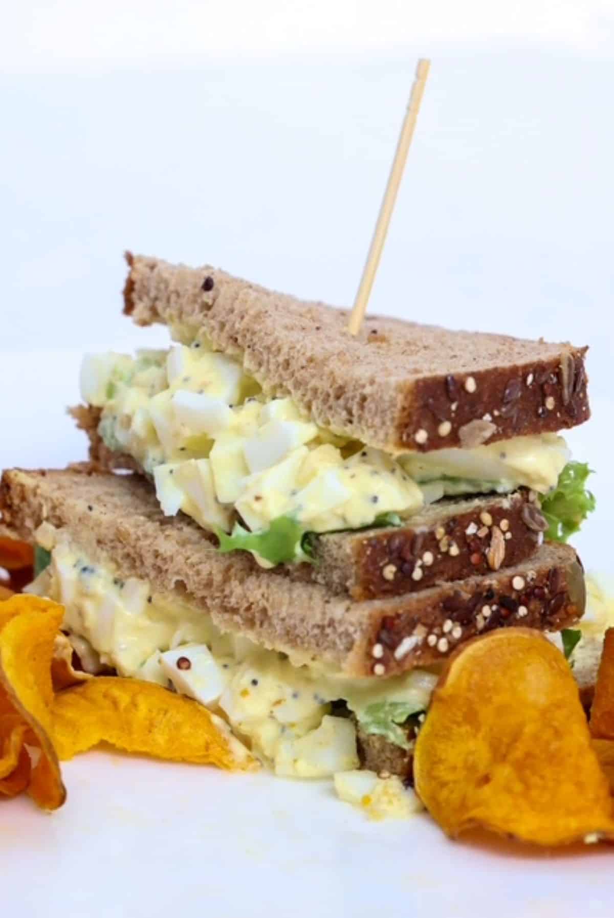 One egg salad sandwich with the halves stacked, on a white plate with a side of sweet potato chips.