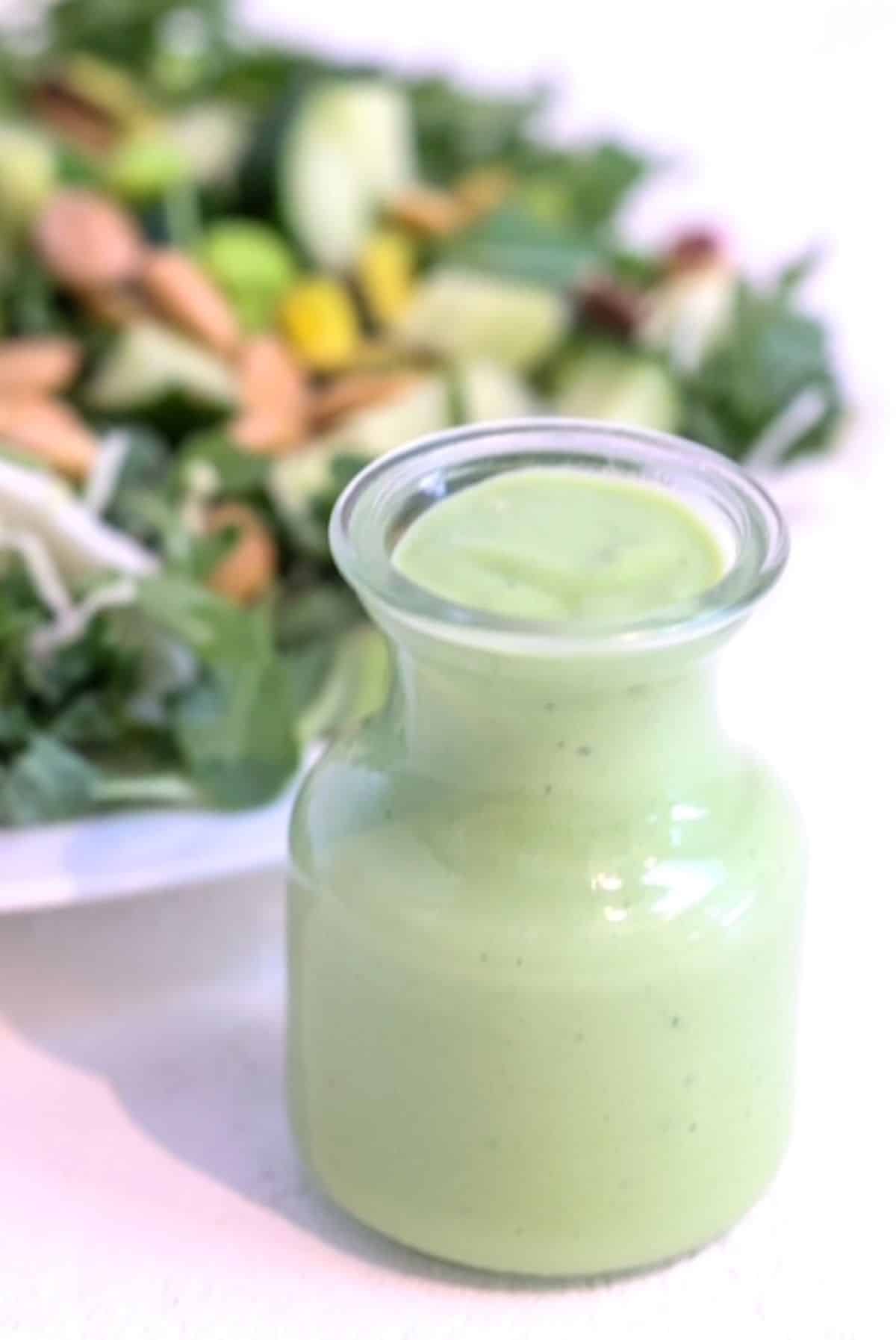 Green Goddess Salad Dressing in a small glass carafe in front of a salad.