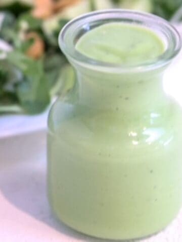 Green goddess dressing in a glass carafe in front of a green salad.