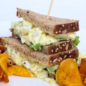 Two halves of an egg salad sandwich stacked, on a plate with sweet potato chips.