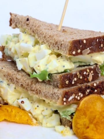 Two halves of an egg salad sandwich stacked, on a plate with sweet potato chips.