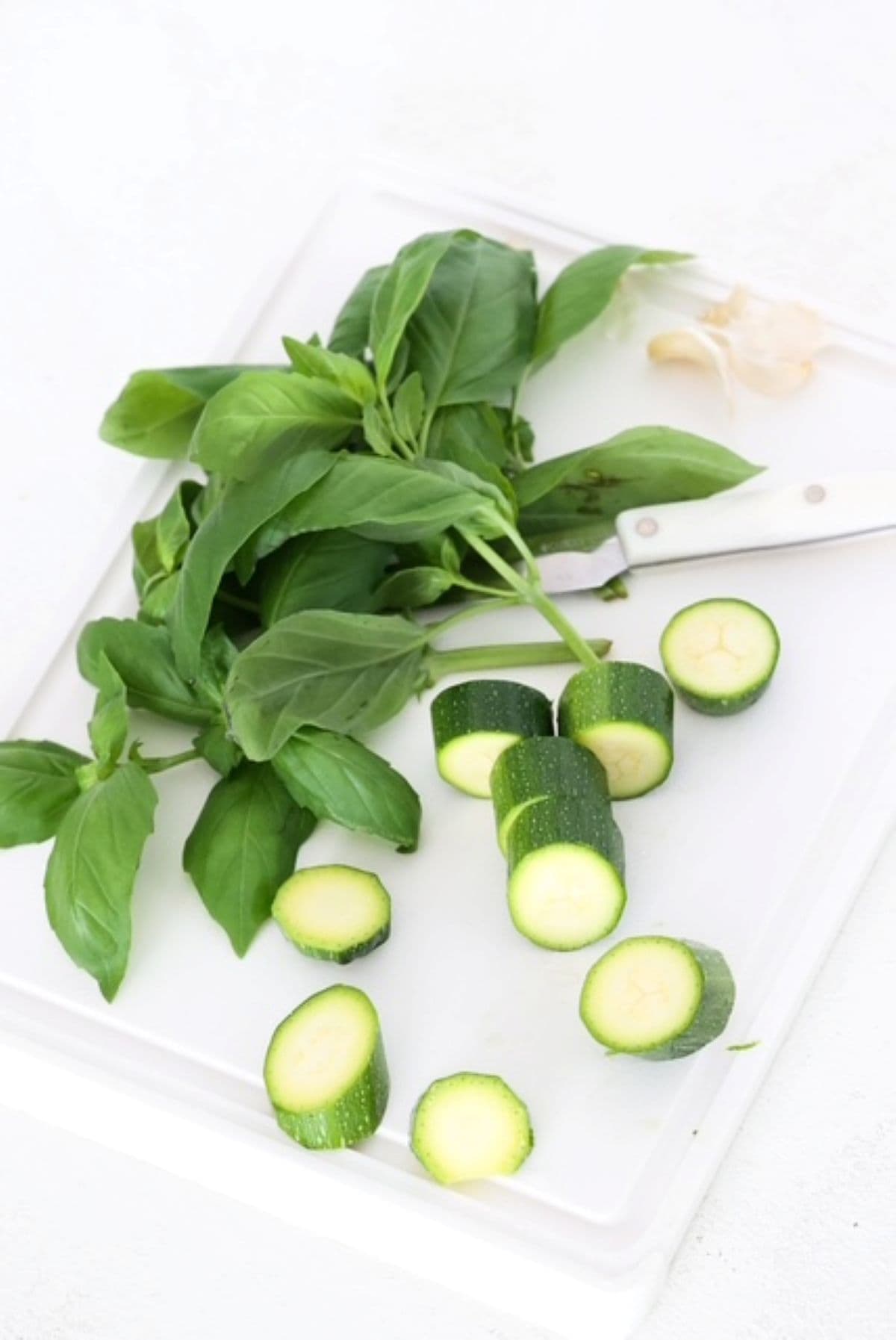Basil, garlic cloves, and sliced zucchini on a white cutting board.